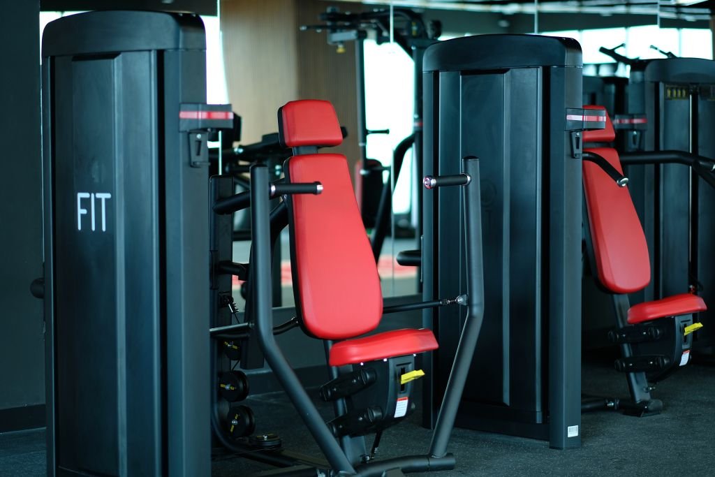 Deretan mesin beban selektor merah dan hitam di area latihan kekuatan Sinergi Fit