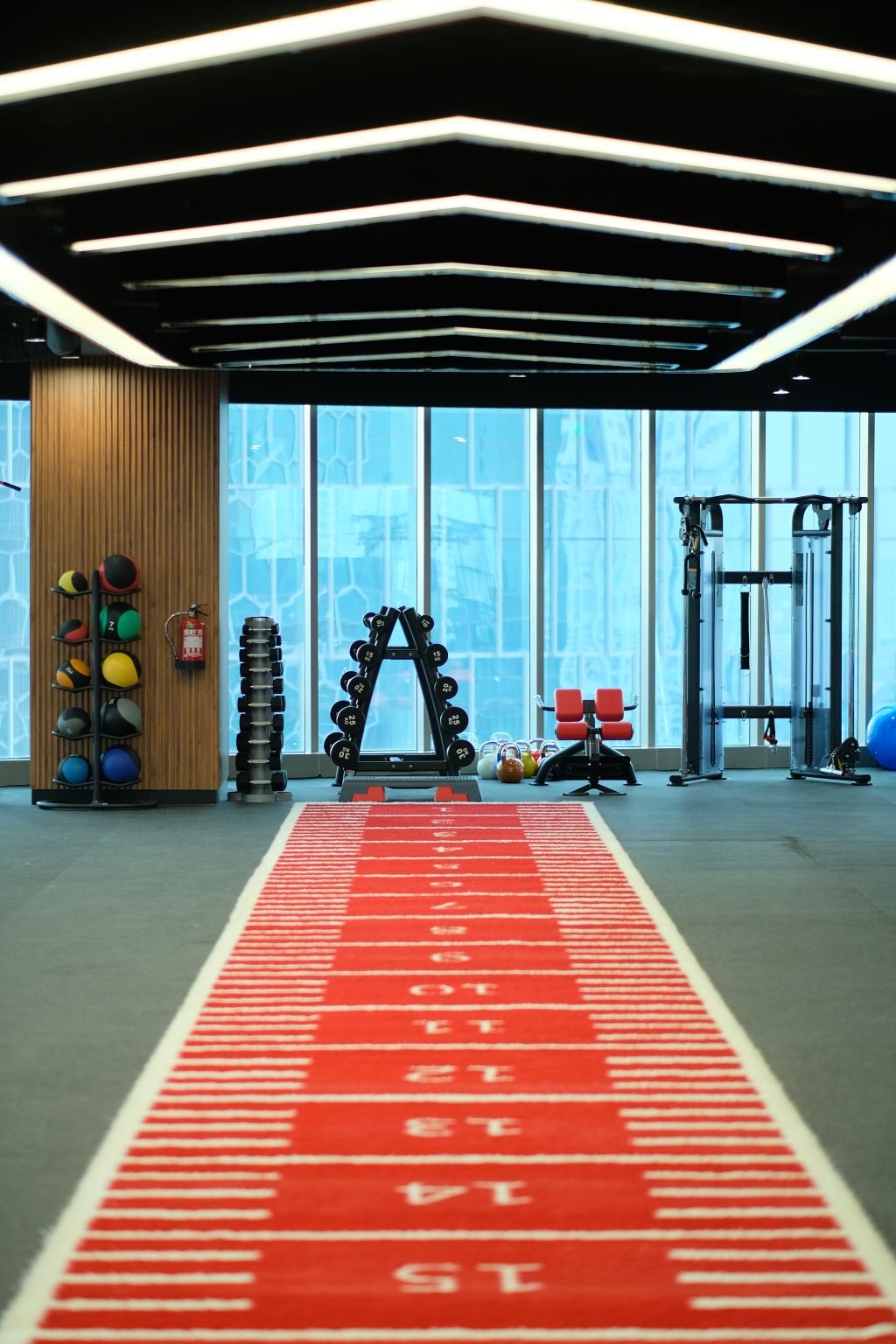 Area latihan fungsional Sinergi Fit dengan lintasan sprint merah, rak medicine ball, dan lampu plafon heksagonal